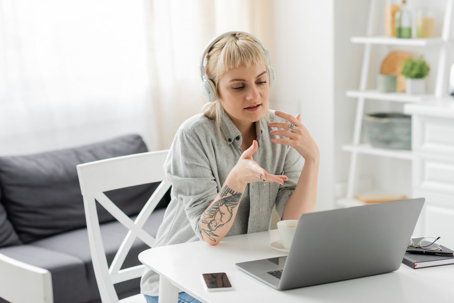 young woman with blonde hair, bangs and tattoo on hand sitting in wireless headphones and using